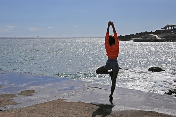 A woman wearing orange top in yoga tree pose near the beach. 
